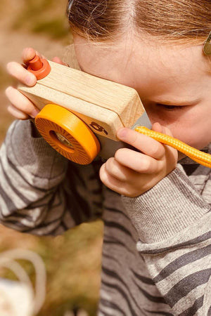 A child holds a wooden toy camera with an orange lens, capturing images during an outdoor adventure. The camera features a natural wood finish and is designed for imaginative play.