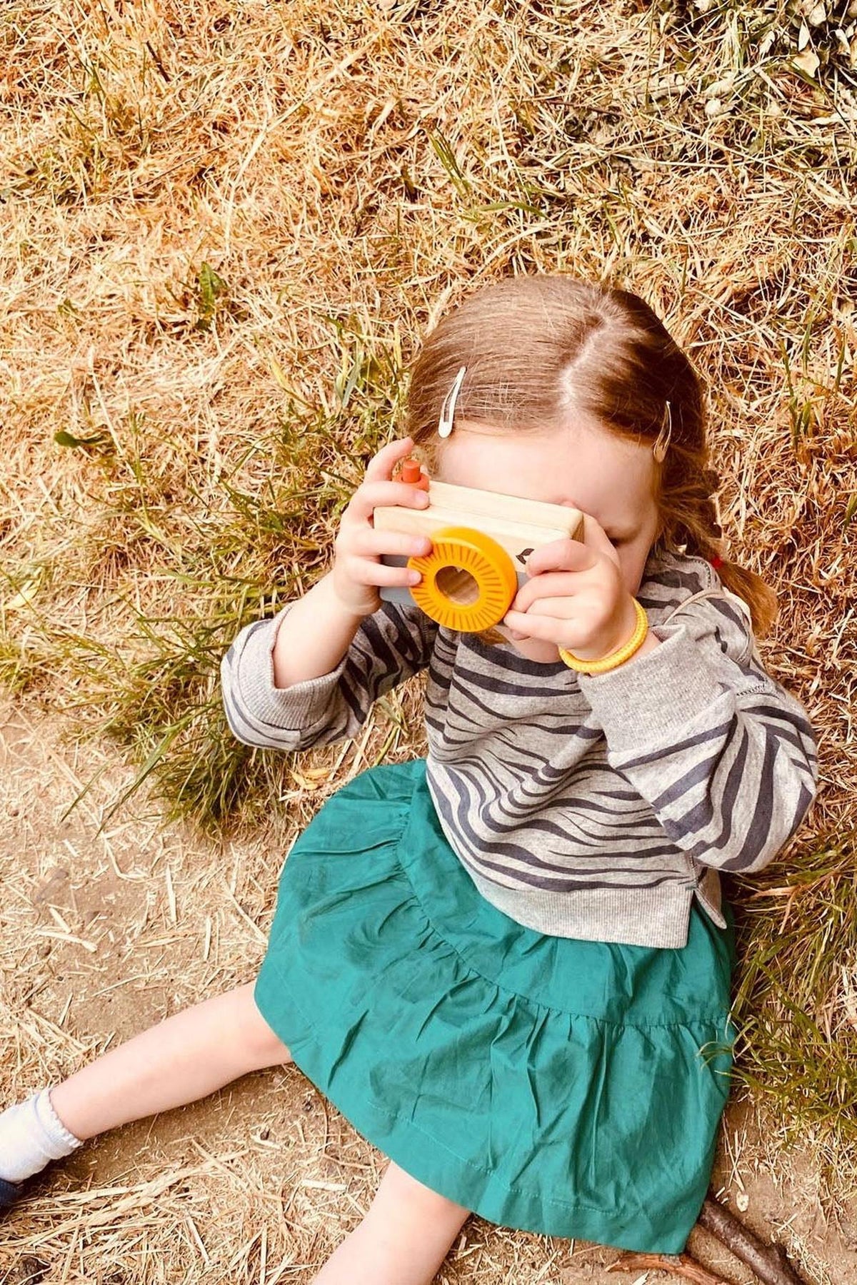 A young girl sits on the ground in a grassy area, wearing a striped sweater and a green skirt, using a camera to capture images of her surroundings.