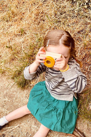 A young girl sits on the ground in a grassy area, wearing a striped sweater and a green skirt, using a camera to capture images of her surroundings.