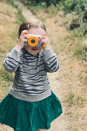 A young child wearing a striped gray sweater and a green skirt holds a bright orange camera, smiling while standing on a grassy path surrounded by greenery.