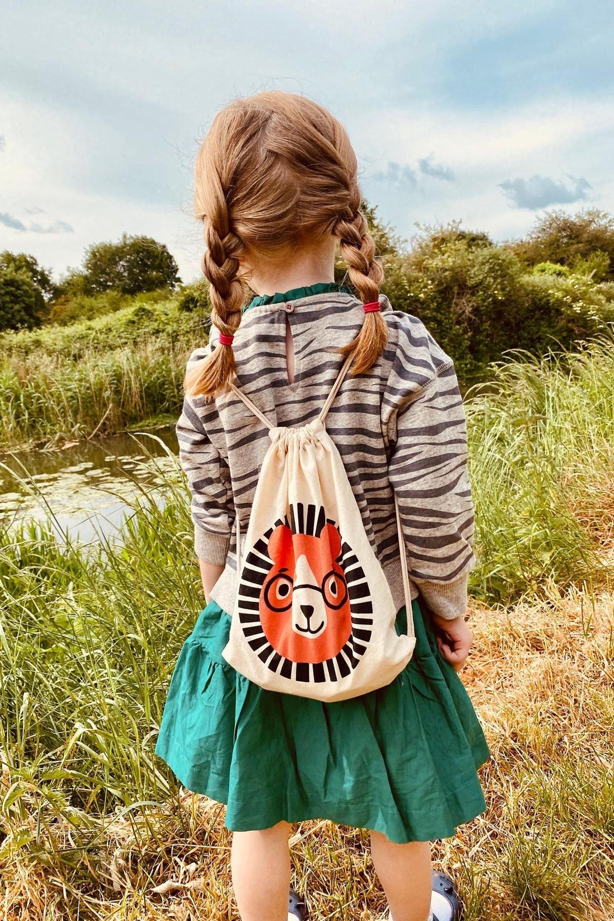 A young girl with braided hair stands by a waterway, wearing a striped shirt and green dress.