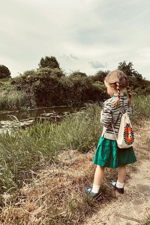 A young girl with braided hair stands by a riverbank, wearing a striped hoodie and green skirt. She holds a drawstring bag featuring a vibrant design, surrounded by lush greenery.