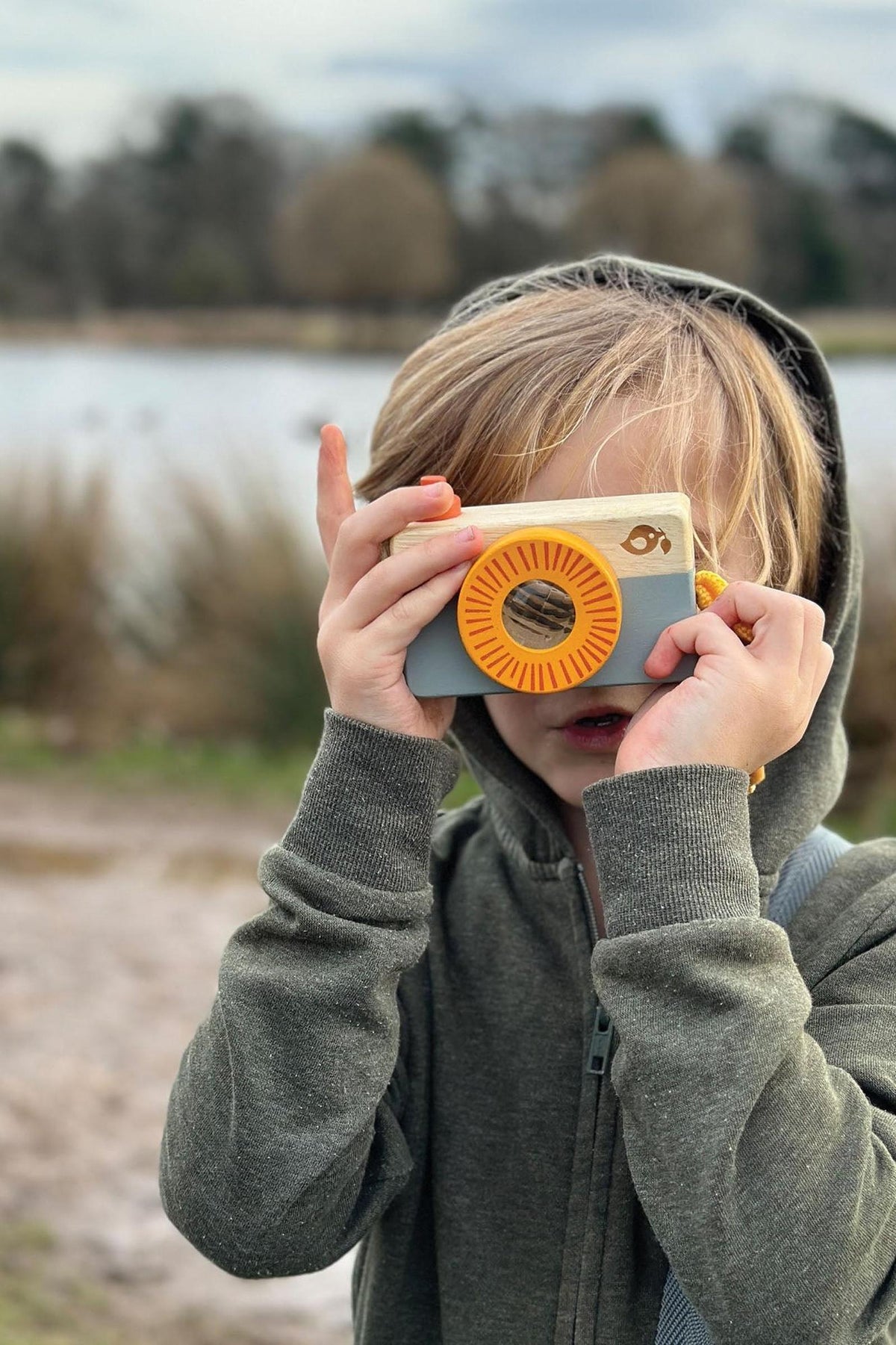 A child wearing a gray hoodie holds a colorful toy camera up to their face, posing playfully near a body of water surrounded by grass and trees.