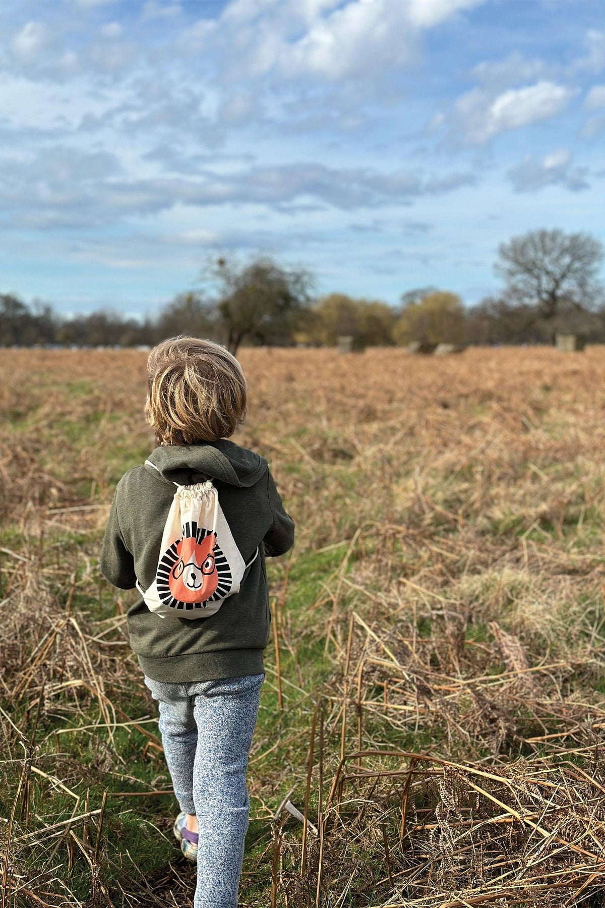 A child walks through a grassy field, wearing a green hoodie and gray pants, with a drawstring bag featuring a playful lion design on their back.