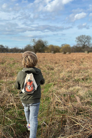 A child walks through a grassy field, wearing a green hoodie and gray pants, with a drawstring bag featuring a playful lion design on their back.