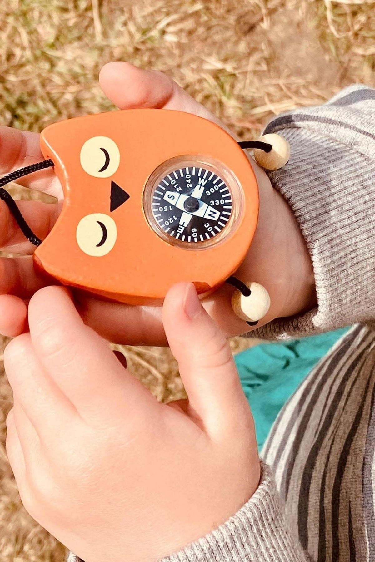 A child holds a whimsical orange owl compass with a black dial, featuring closed eyes and a beak. The background shows grassy terrain, emphasizing outdoor exploration and adventure.