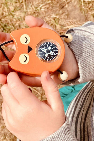 A child holds a whimsical orange owl compass with a black dial, featuring closed eyes and a beak. The background shows grassy terrain, emphasizing outdoor exploration and adventure.