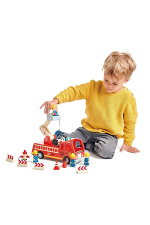 A young child wearing a yellow sweater plays with a red wooden fire engine, surrounded by traffic cones and figures.
