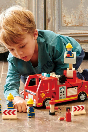 A young child plays with a vibrant red wooden fire engine toy, featuring firefighter figures and accessories on a textured.