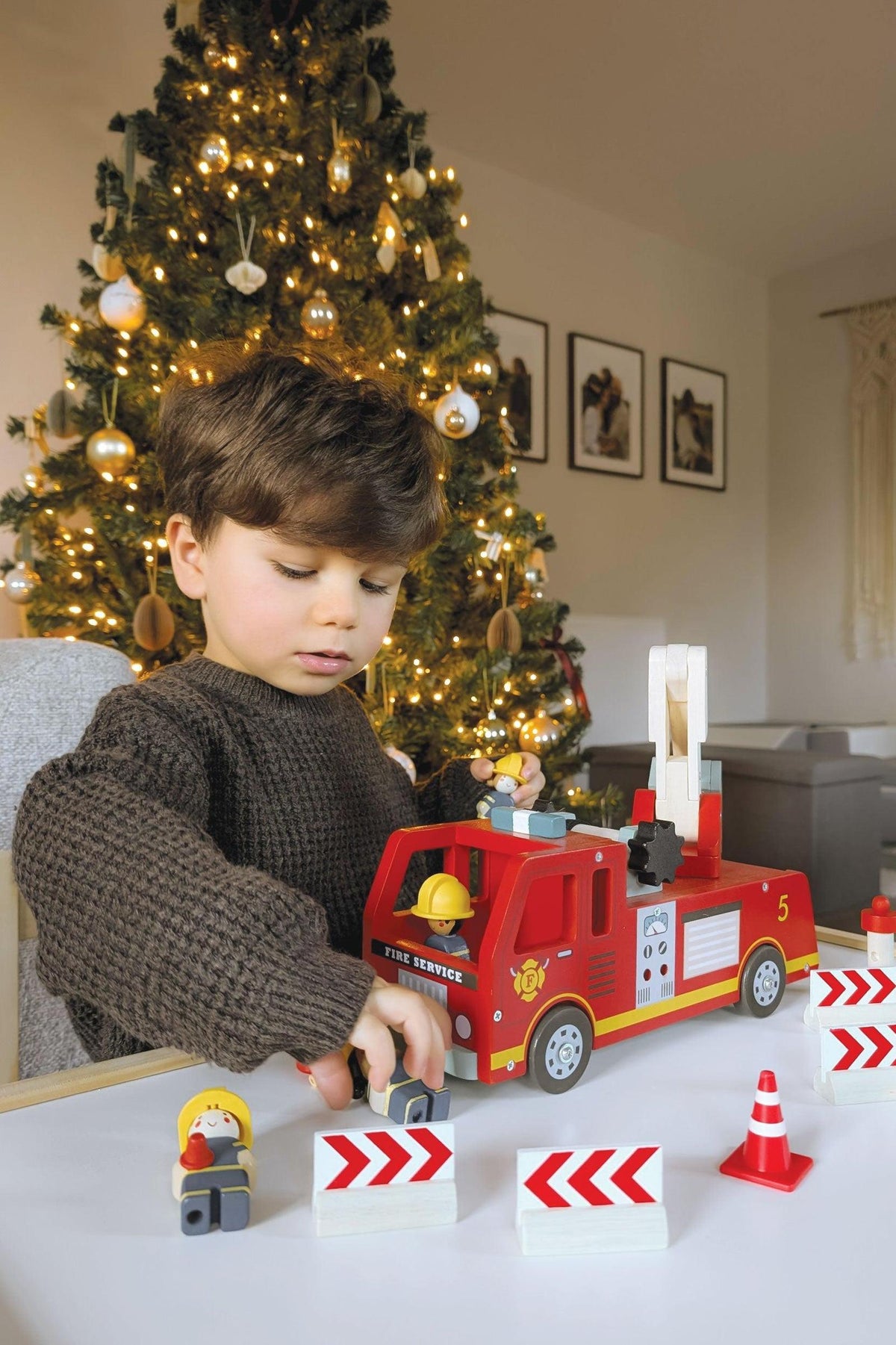 A young child plays with a vibrant red wooden fire engine toy, surrounded by festive holiday decorations and a Christmas.
