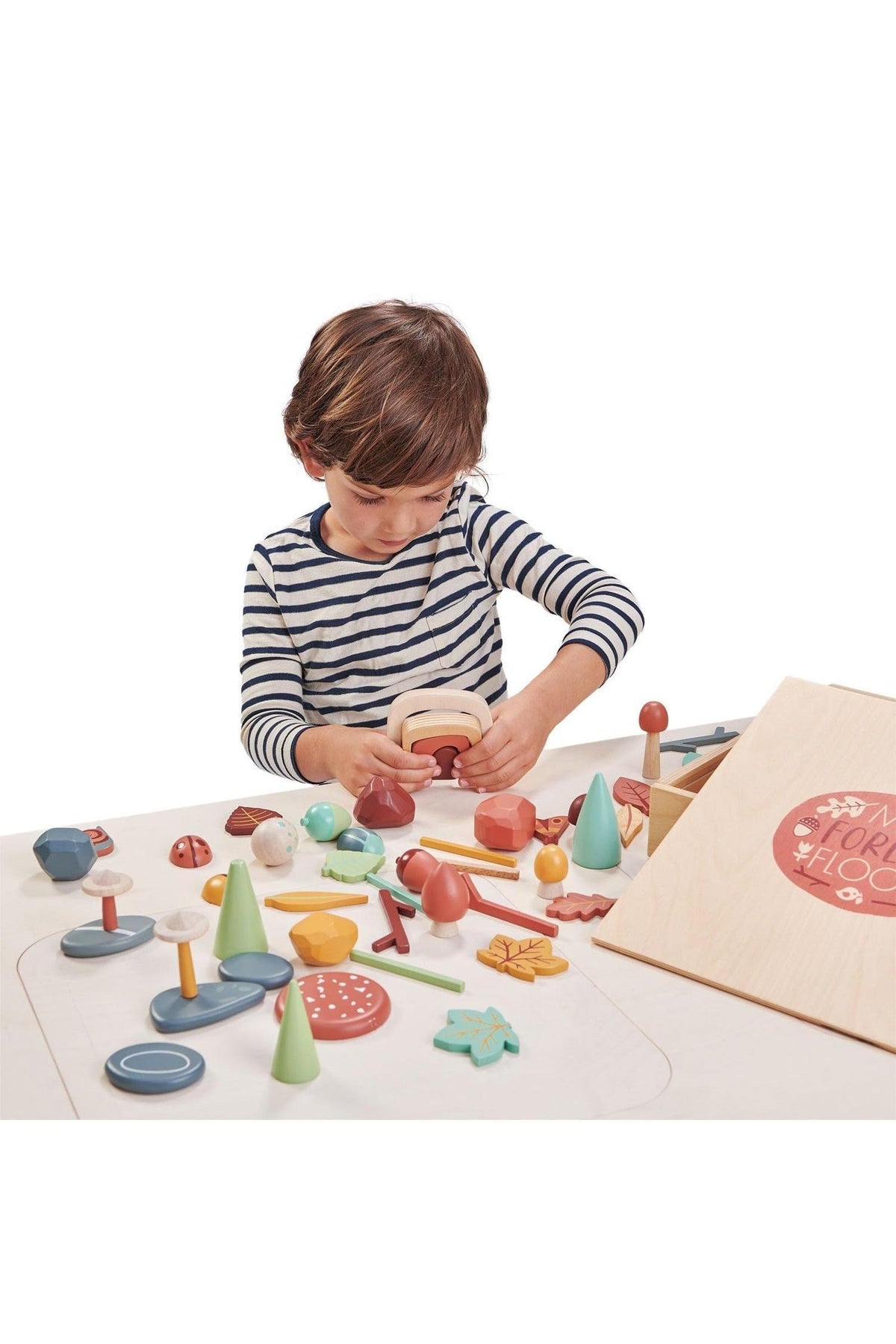 A young child in a striped shirt interacts with colorful wooden pieces from the Tender Leaf My Forest Floor toy set, exploring shapes and textures on a light-colored play surface.