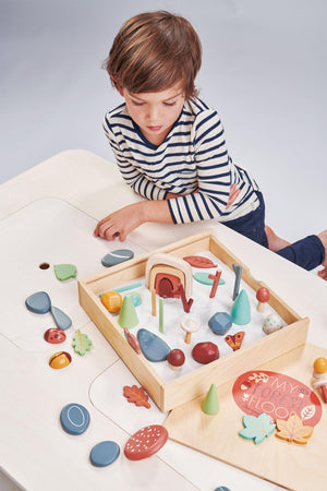 A child examines a wooden sorting tray filled with colorful woodland-themed pieces, including leaves and mushrooms, set against a light-colored table. The toy encourages imaginative play and creativity.
