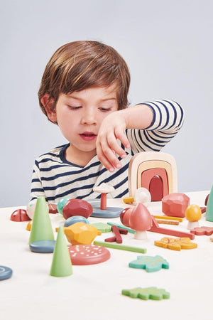 A young child with brown hair wearing a striped shirt engages with colorful, wooden toy shapes, including trees, mushrooms, and geometric figures, on a light-colored table.
