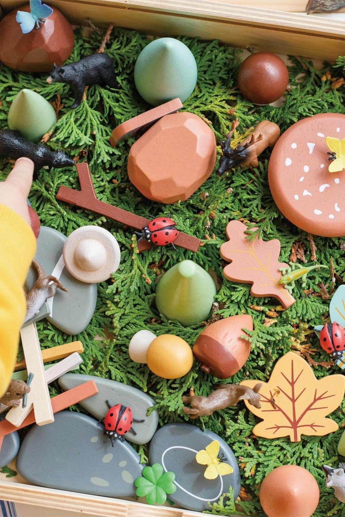 A colorful wooden play tray filled with various natural-themed figurines, stones, and leaves is arranged on a bed of green foliage. The items encourage imaginative play and creativity for children.