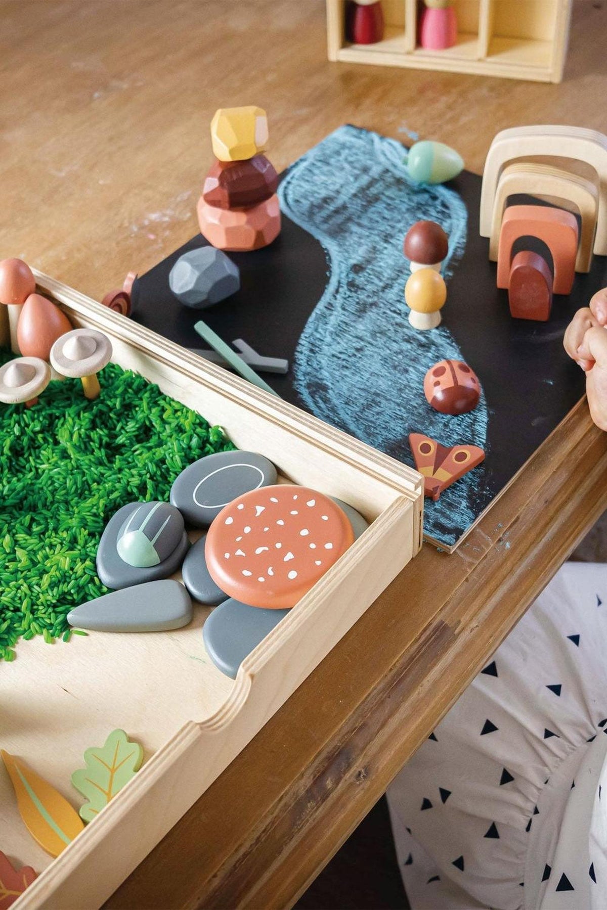 A wooden tinker tray filled with colorful, eco-friendly shapes, including rocks, leaves, and mushrooms, sits on a wooden table. The reversible lid features a chalkboard for creative play.