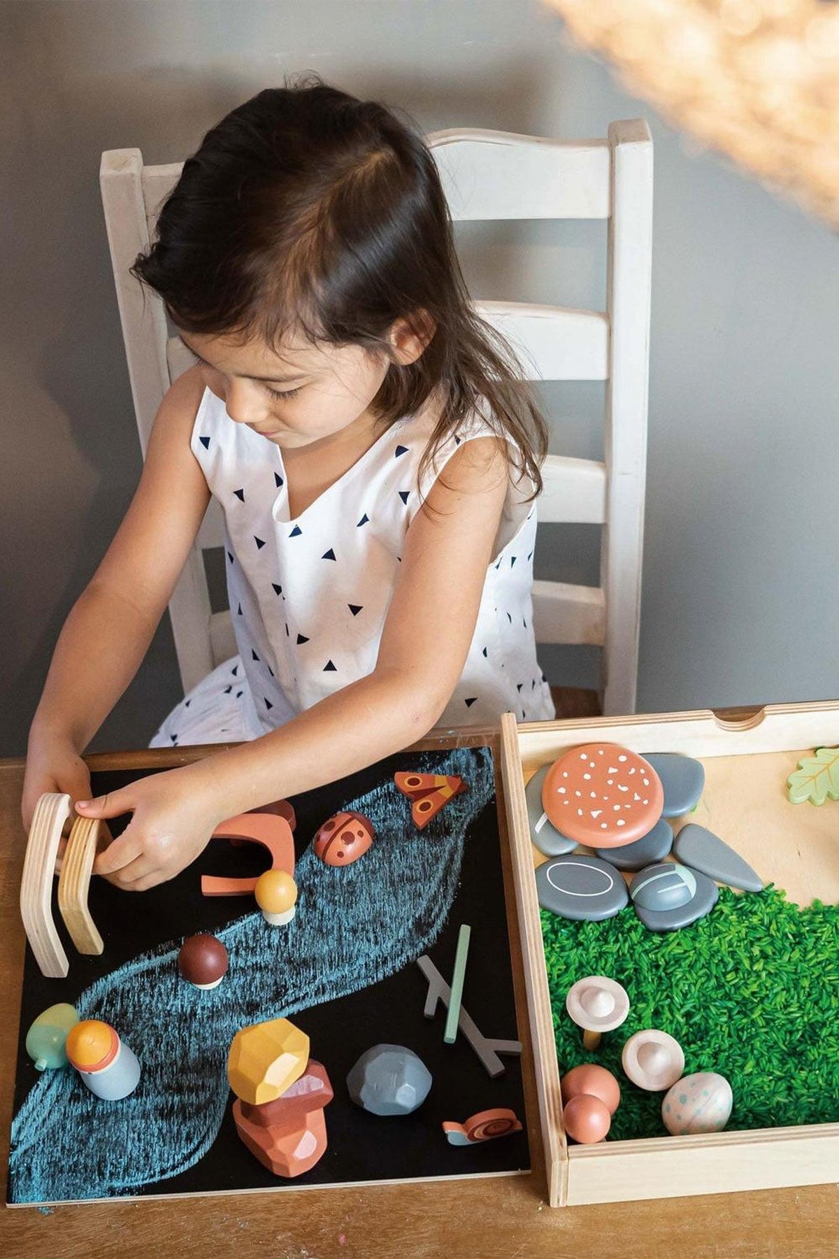 A young child plays with colorful wooden toys on a reversible tray, featuring a chalkboard surface and compartments filled with vibrant, nature-themed pieces. The setting is bright and inviting.
