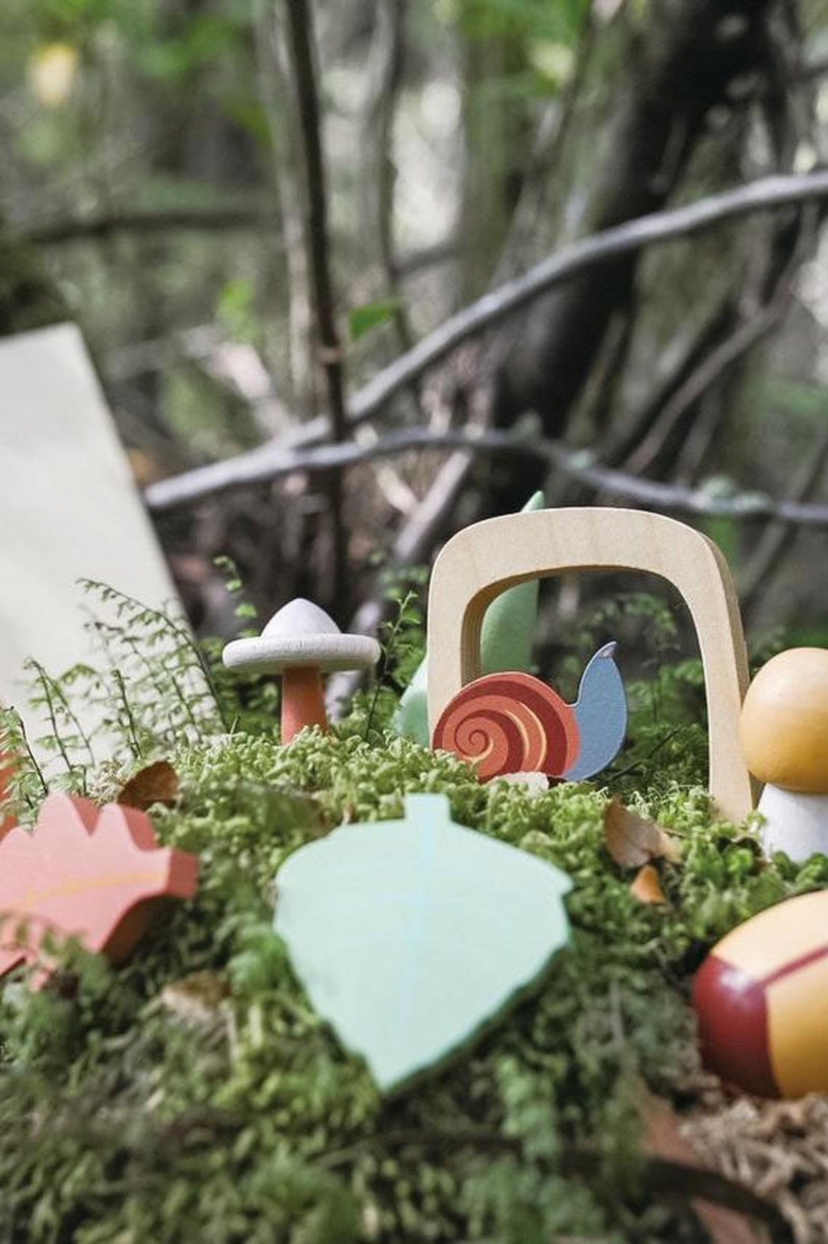 A colorful arrangement of wooden toys, including a snail, mushroom, and leaf, is nestled among lush green moss in a forest setting, showcasing organic and sustainable play features.