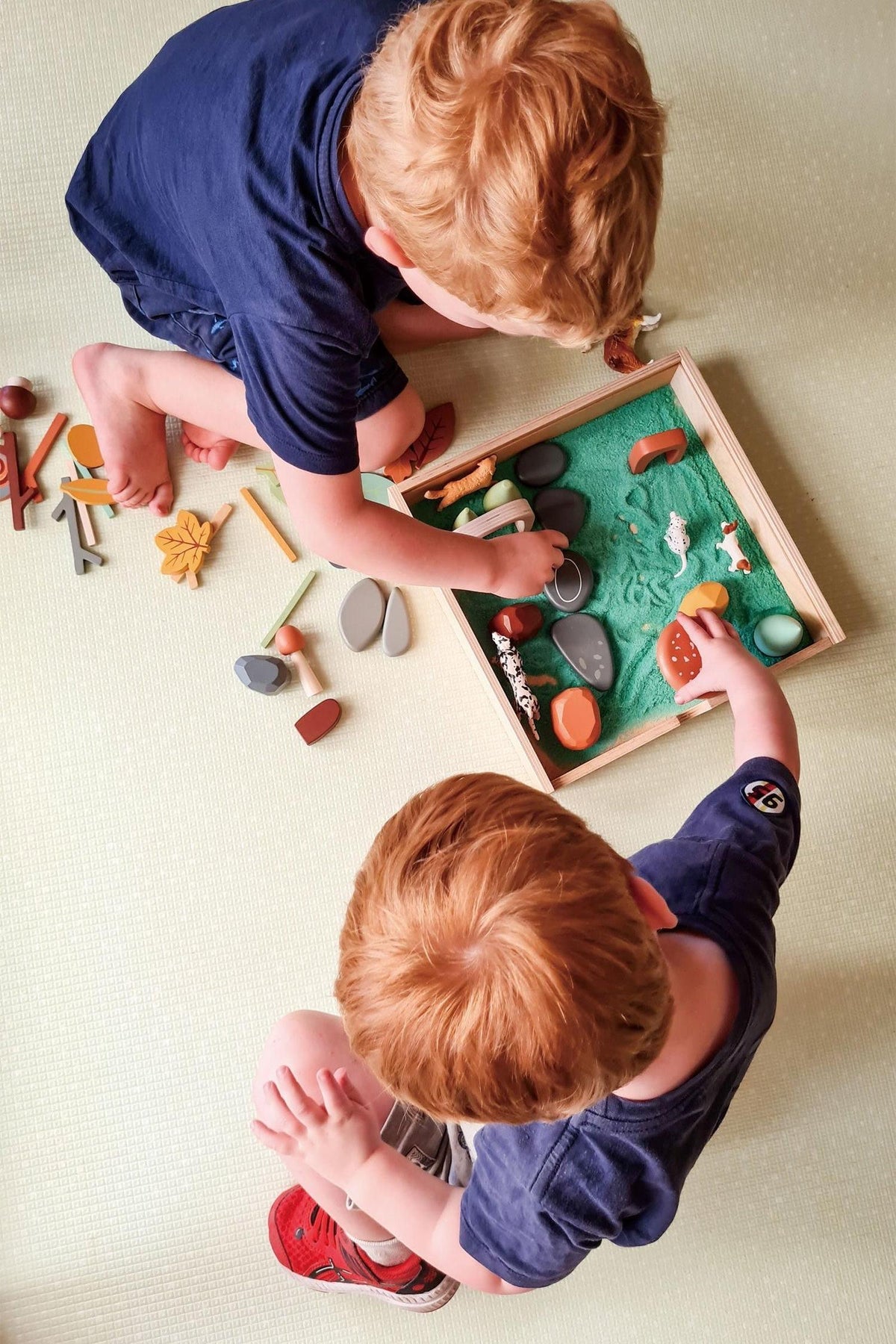Two young boys play together with a wooden tinker tray filled with colorful, natural elements, exploring their creativity in a bright, indoor setting.