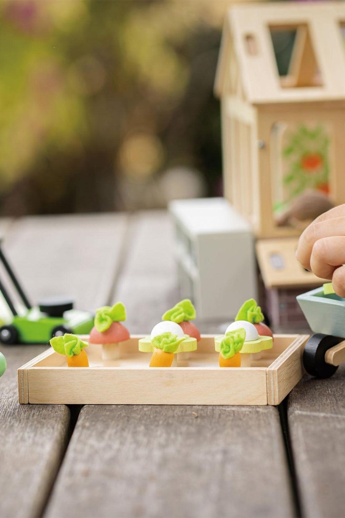 A wooden tray holds colorful toy vegetables, including carrots and lettuce, ideal for imaginative play. The scene features a child’s hand engaging with the Tender Leaf Wooden Toys garden set.