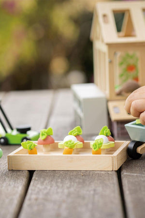 A wooden tray holds colorful toy vegetables, including carrots and lettuce, ideal for imaginative play. The scene features a child’s hand engaging with the Tender Leaf Wooden Toys garden set.