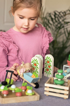 A young girl in a pink shirt plays with a wooden toy garden set, featuring colorful vegetables, a wheelbarrow, and decorative garden panels. Sustainable materials enhance imaginative play.