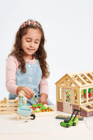 A young girl wearing a pink long-sleeve shirt and denim overalls waters colorful wooden vegetables beside a detailed wooden greenhouse, part of the Tender Leaf Wooden Toys Garden Set.