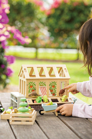 A child interacts with a wooden greenhouse playset, featuring vibrant plants and gardening tools, set against a backdrop of blooming flowers. The eco-friendly design encourages imaginative outdoor play.