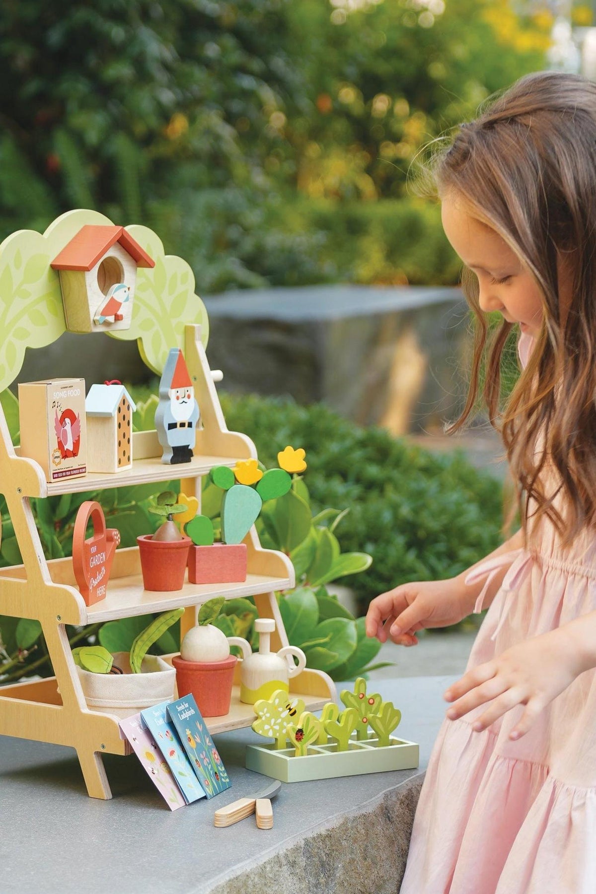 A child in a pink dress interacts with a colorful wooden garden center, featuring various accessories like a birdhouse, garden tools, and potted plants, set against a natural outdoor backdrop.
