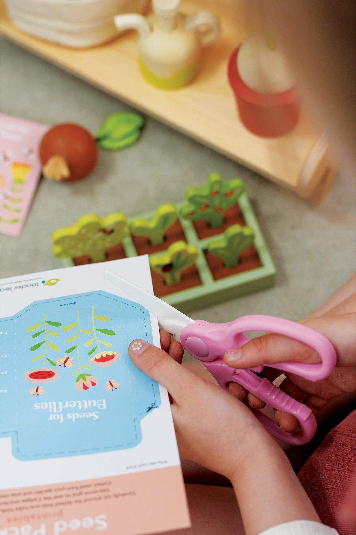 A child uses pink scissors to cut a colorful seed packet illustration while seated on a soft surface. Nearby, vibrant wooden plants and a gardening tray are visible.