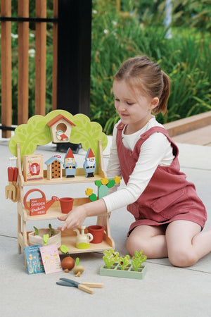 A young girl in a pink romper interacts with a colorful wooden garden center toy, featuring pots, tools, and playful accessories, set against a lush green backdrop.