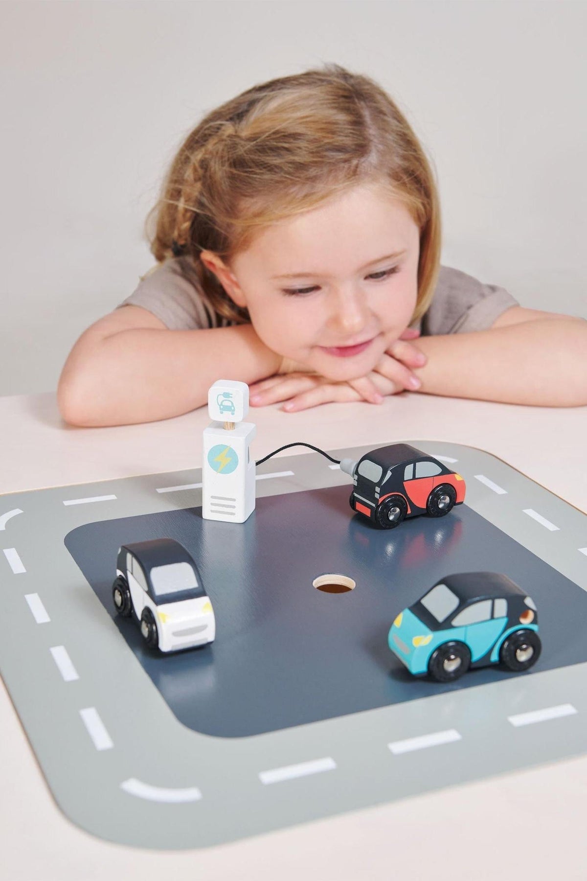 A young girl watches intently as three colorful wooden electric cars are parked around a charging station on a play mat, showcasing sustainable, imaginative play.