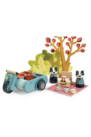A charming wooden picnic set features two badger characters beside a blue motorbike and sidecar, set against a colorful tree. The scene includes a picnic blanket with food items.