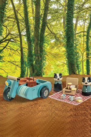 A charming wooden motorbike and sidecar in blue, accompanied by two playful badger figures, are set up for a picnic on a red-checkered blanket among lush, green trees.