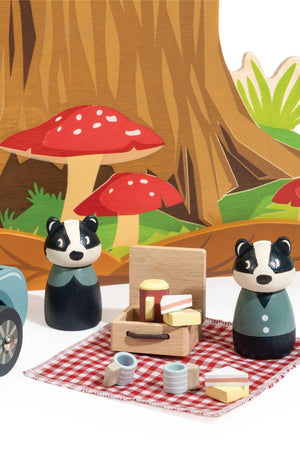 A colorful wooden play set featuring two badger figurines, a picnic basket, and a checkered blanket, set against a woodland backdrop with mushrooms and trees.