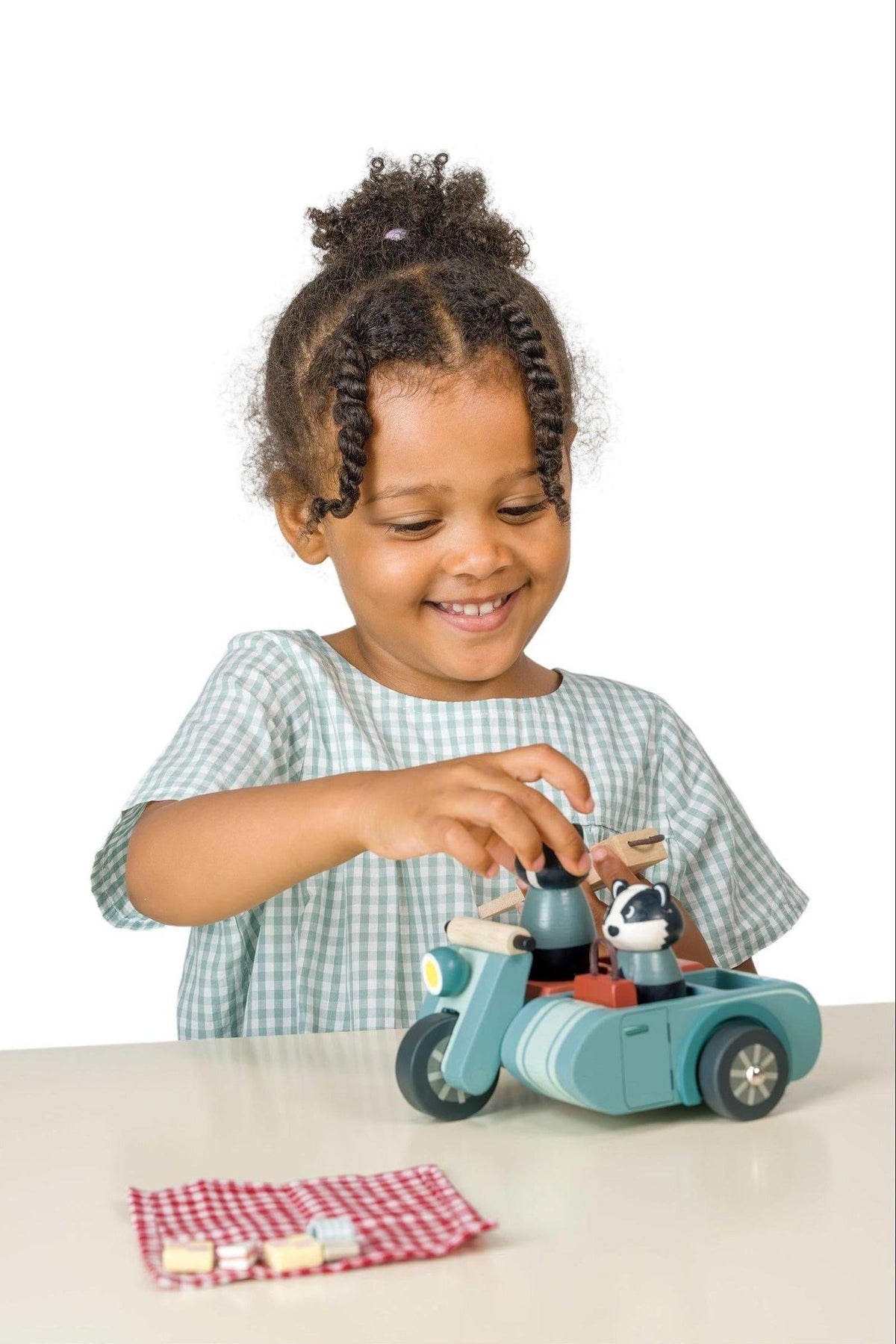 A child smiles while playing with a wooden motorbike toy featuring animal figures. The set includes a picnic blanket with food items, highlighting eco-friendly and sustainable play features.