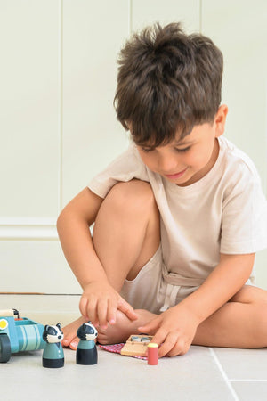 A young child sits on the floor, playing with two wooden badger figures and a picnic set. The scene features a light, airy setting with neutral-colored walls.