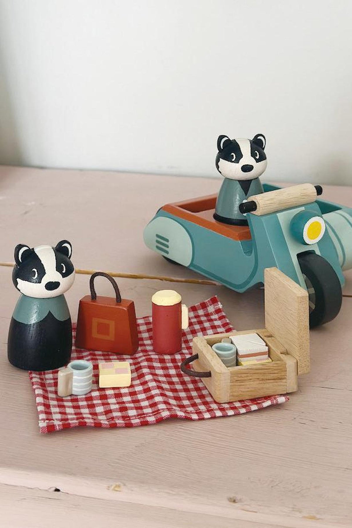 A charming wooden motorbike in teal, accompanied by two badger figures, sits next to a picnic setup on a red and white checkered blanket, featuring a basket and food items.