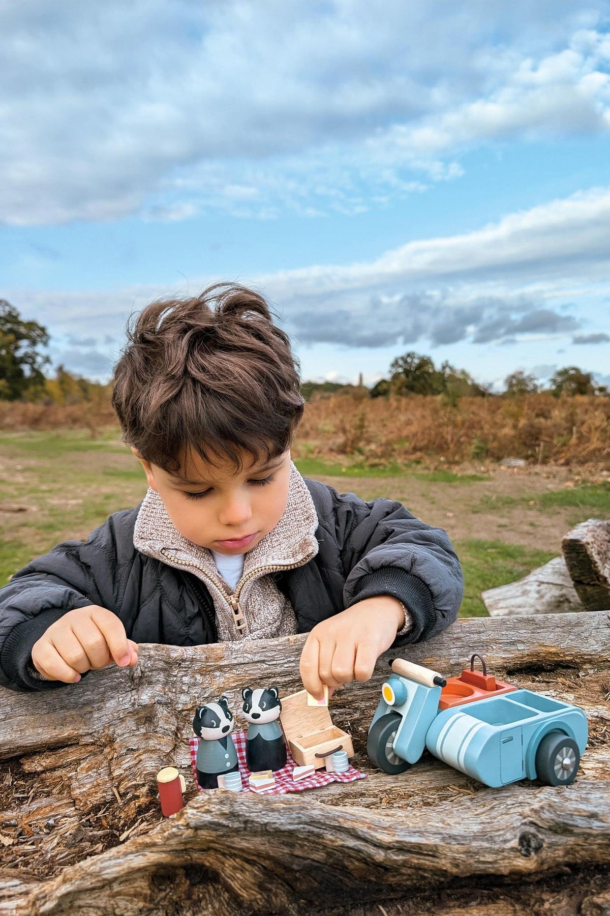 A young boy plays outdoors with a Tender Leaf wooden toy set featuring two animal figures, a picnic basket.