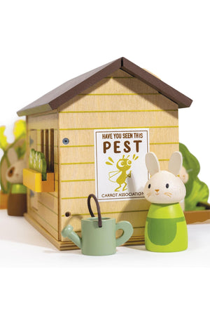 A wooden playset features a cozy shed with a Have You Seen This Pest sign, a bunny character, and a green watering can. The colorful design encourages imaginative gardening play.