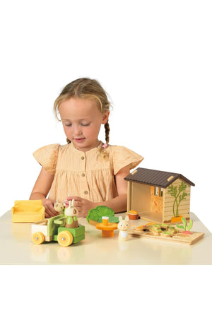 A young girl plays with the Tender Leaf Wooden Toys Bunnys Carrot Patch set, featuring a wooden shed, a toy tractor, and vegetable pieces, all designed for imaginative, eco-friendly play.