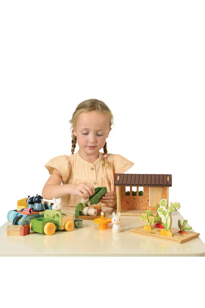 A young girl dressed in a light peach top plays with Tender Leaf Wooden Toys, Bunny’s Carrot Patch, featuring a wooden shed, colorful vegetables, and adorable bunny figures.