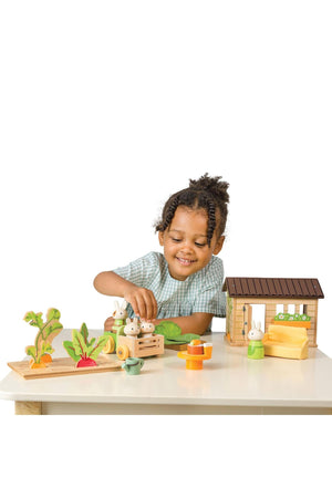 A child smiles while playing with the Tender Leaf Wooden Toys Bunnys Carrot Patch set, featuring wooden rabbits, vibrant vegetables, and a cozy shed, fostering imaginative gardening play.