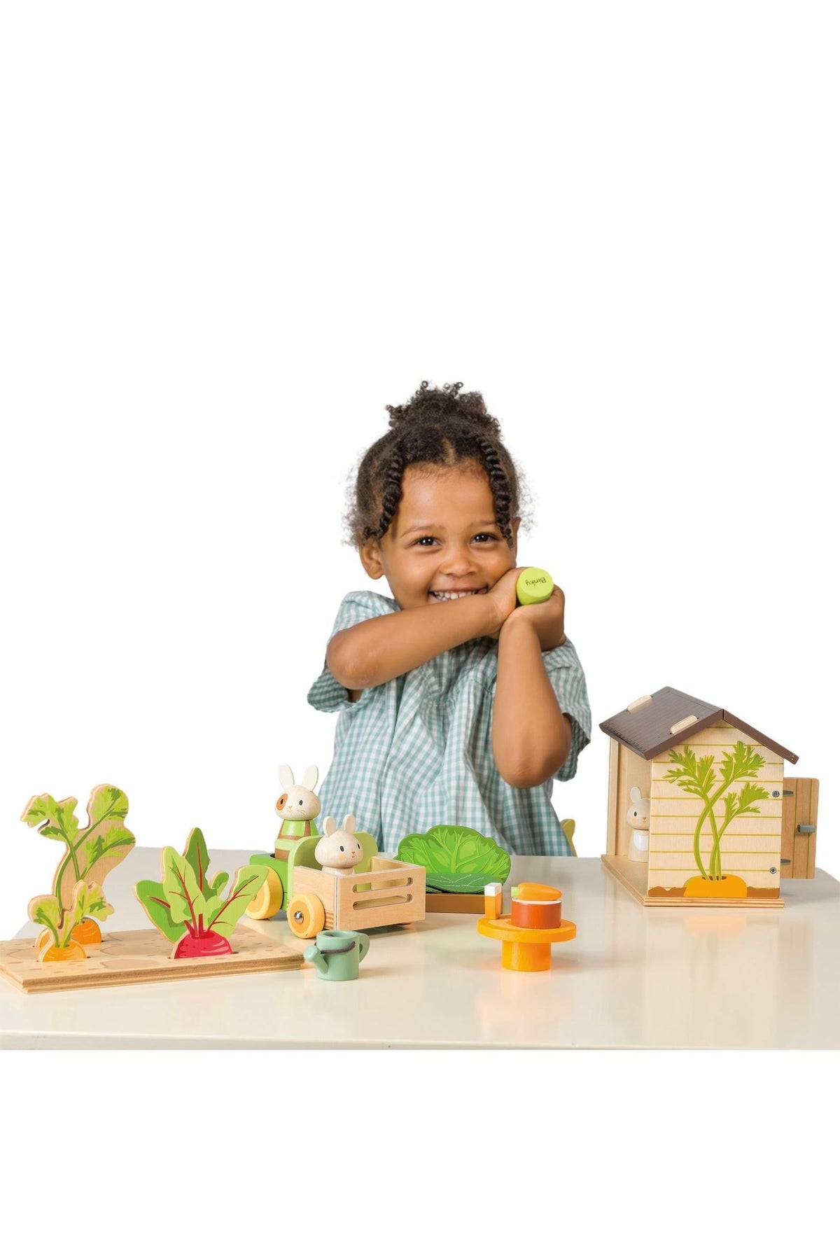 A smiling child in a green checkered dress holds a toy vegetable while surrounded by wooden gardening toys, including a shed and various colorful vegetables, promoting imaginative play.