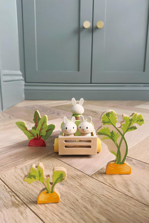 Two wooden bunny figurines sit in a yellow toy cart surrounded by colorful vegetable cutouts, including carrots and beets, on a light wooden floor, creating an inviting play scene.