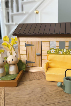 A cozy wooden shed with a brown roof features colorful flower designs, surrounded by two wooden rabbit figures and a yellow couch, perfect for imaginative play. Ideal for young children.