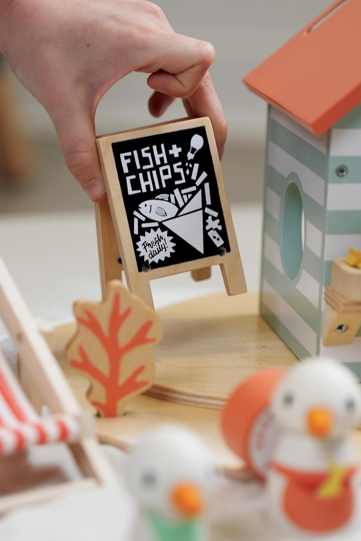 A hand holds a small wooden sign displaying Fish and Chips near a beach hut toy, crafted from sustainable materials.