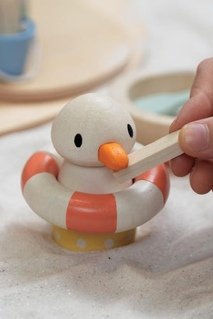 A childs hand holds a wooden paddle near a painted seagull figurine in a lifebuoy, set on a sandy surface.