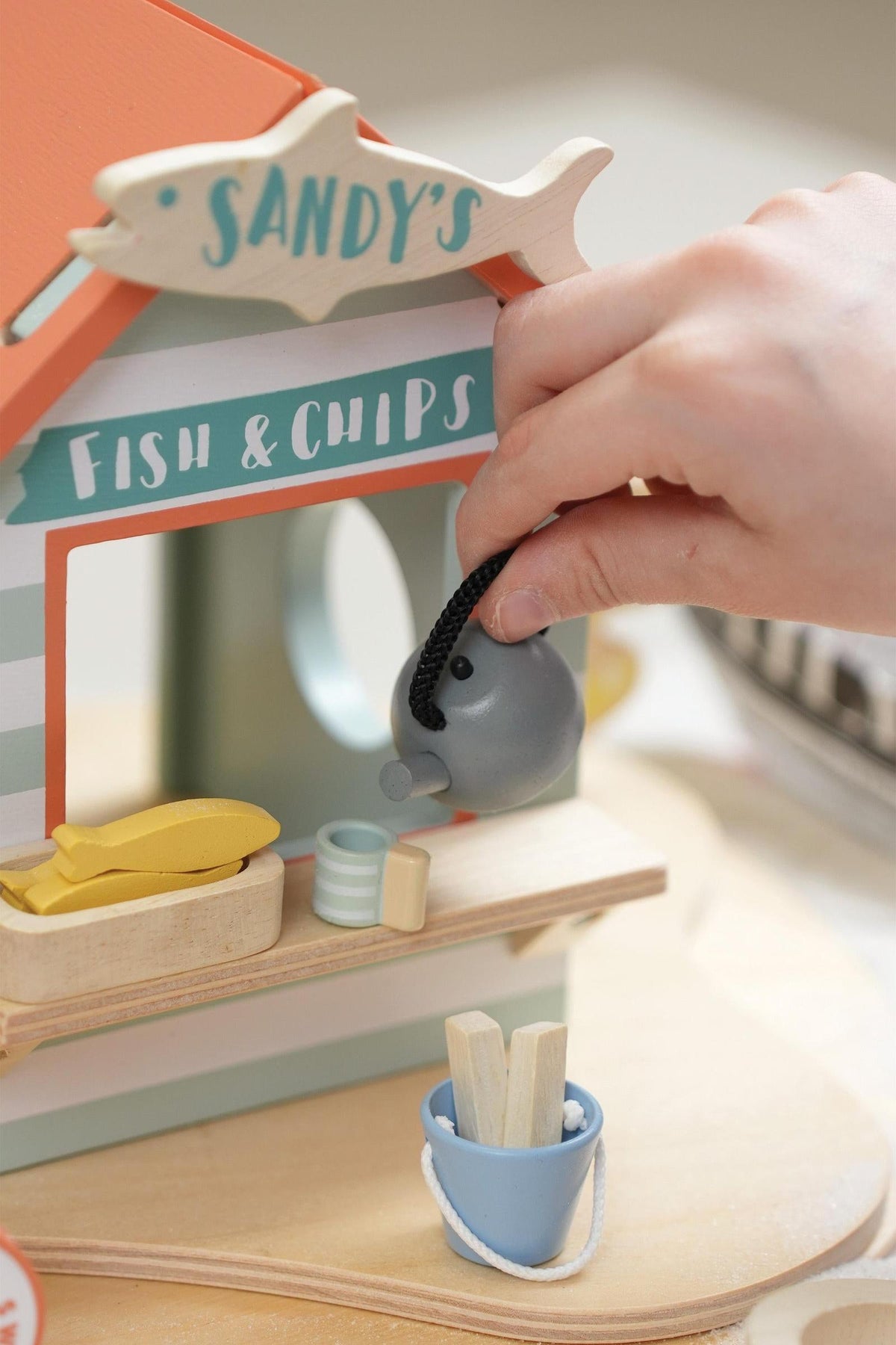 A child’s hand holds a gray wooden elephant near a beach hut labeled Sandys Fish & Chips, featuring colorful accents.