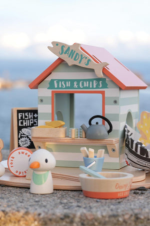A colorful wooden beach hut playset features a fish and chips stall with a kettle, utensils, and a wooden seagull.