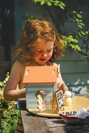 A young child with curly red hair interacts with a wooden beach hut toy, featuring a crab and food accessories.